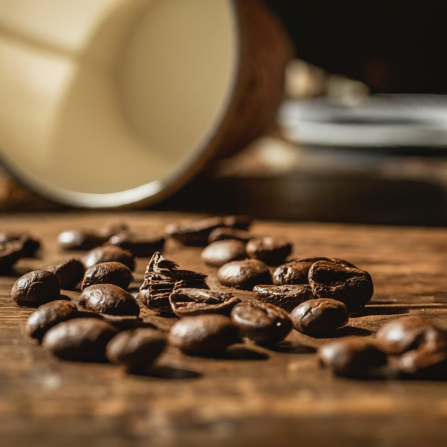 Close-up view of scattered freshly roasted coffee beans on a wooden surface, with a blurred white ceramic cup in the background.